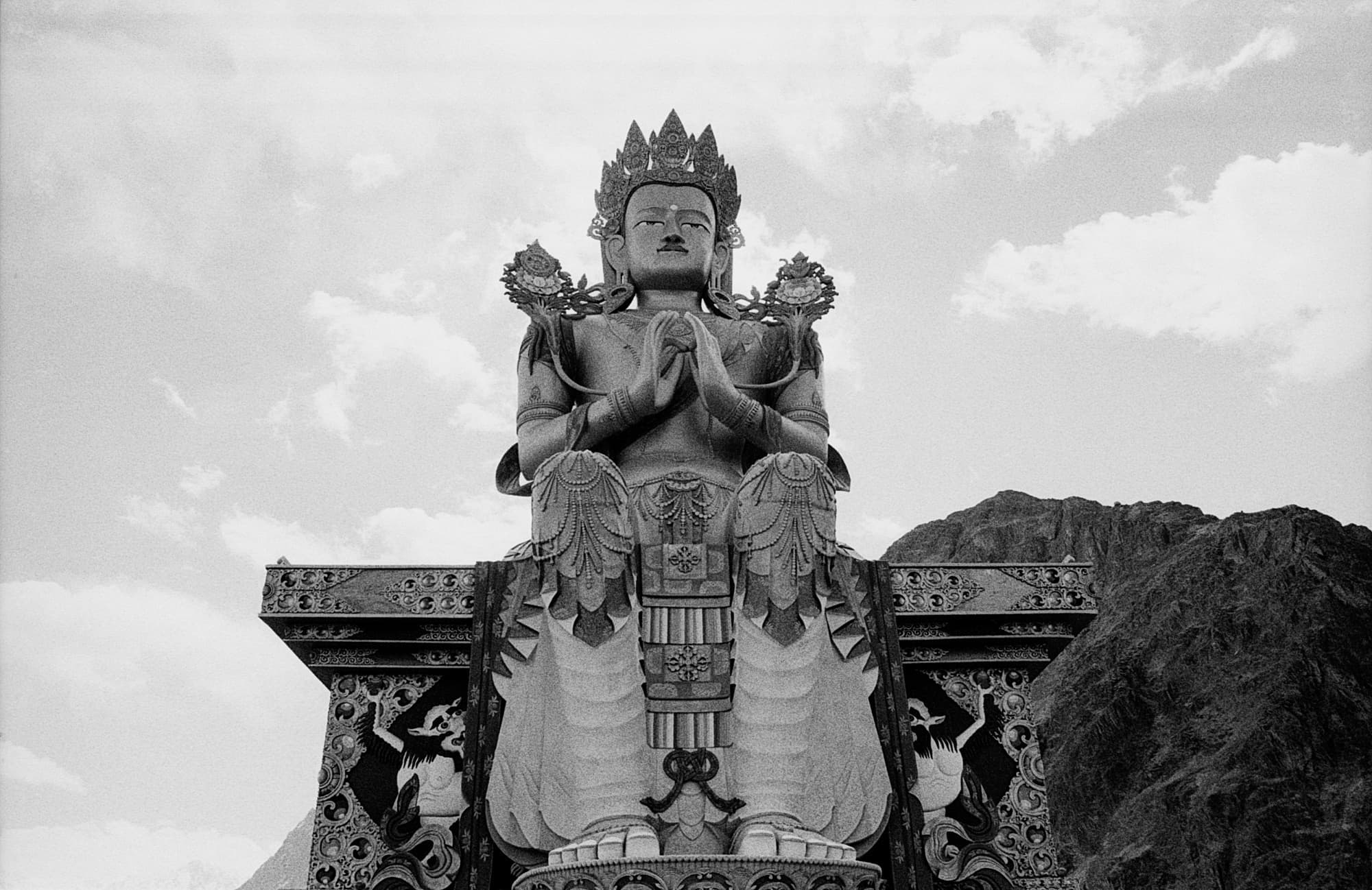 A low-angle view of a large Buddhist statue with hands folded in prayer, set against clouds and mountains