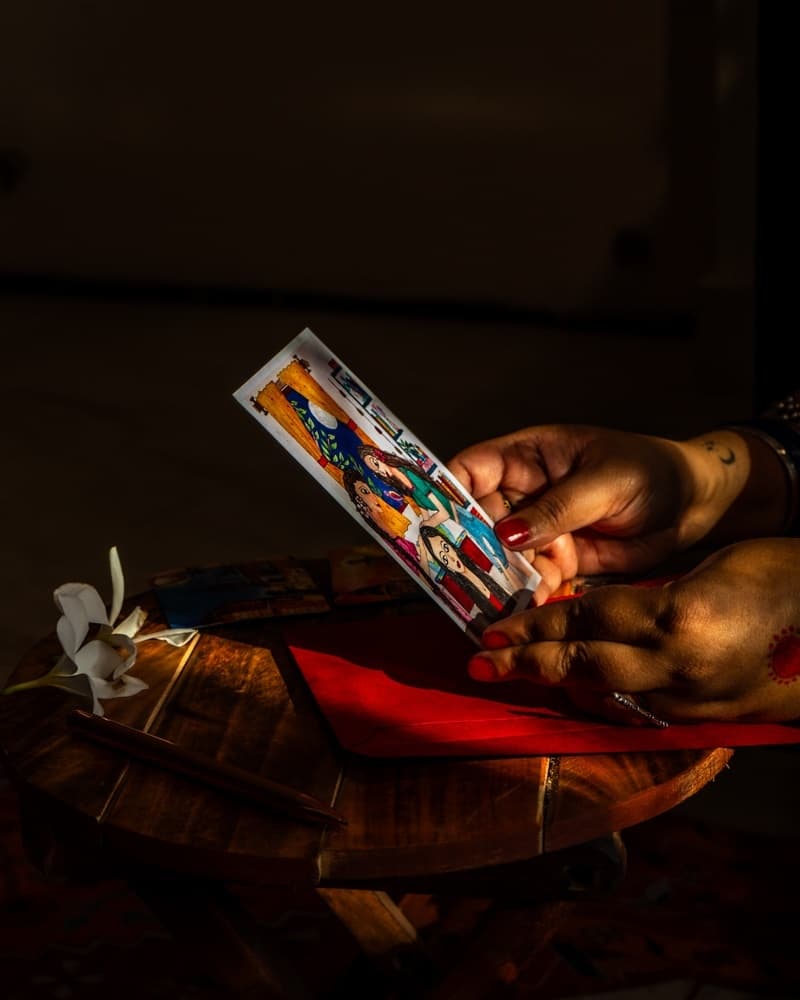 An illustrated bookmark held over a wooden side table scattered with coasters and frangipani flowers