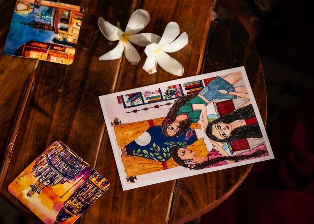 An illustrated postcard resting on a wooden table beside coasters and frangipani flowers