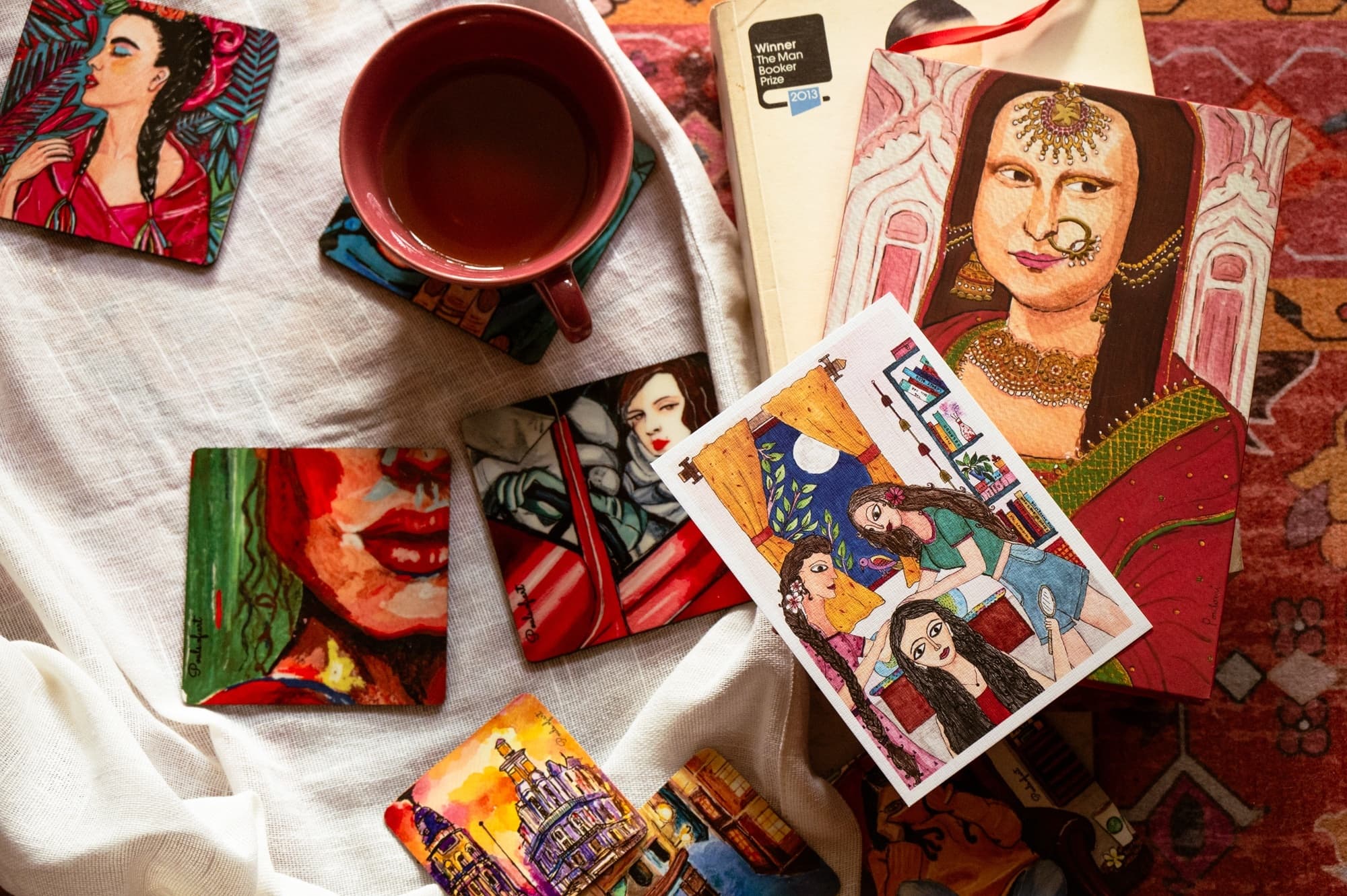A flat lay of illustrated coasters, postcards, and a journal beside a cup of tea and a book