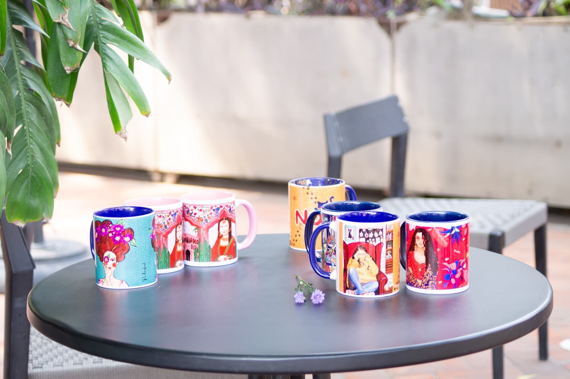 Six illustrated mugs arranged on an outdoor table beside potted plants and flowers
