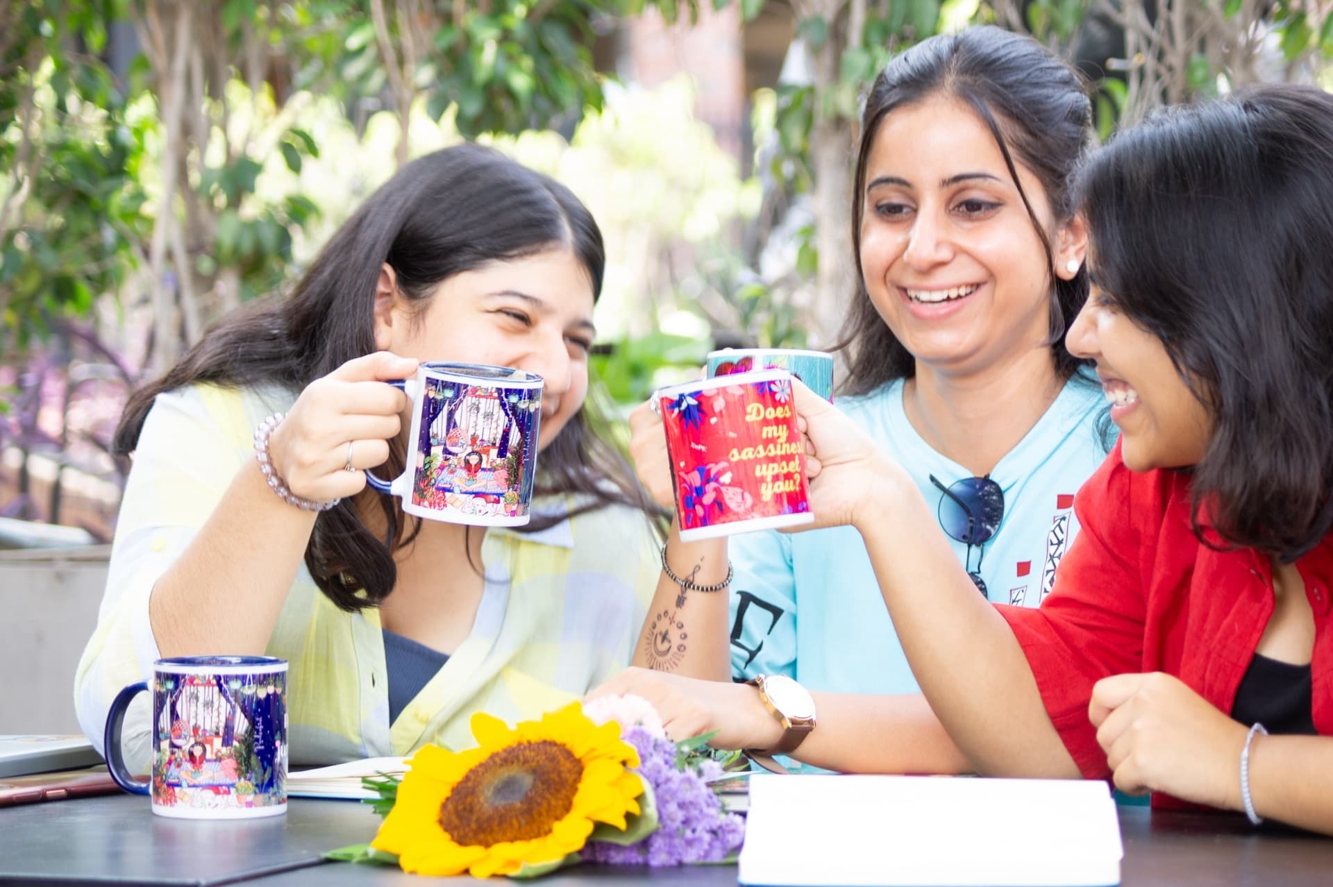 Three women laughing together while two hold up illustrated mugs, with a sunflower and lavender on the table