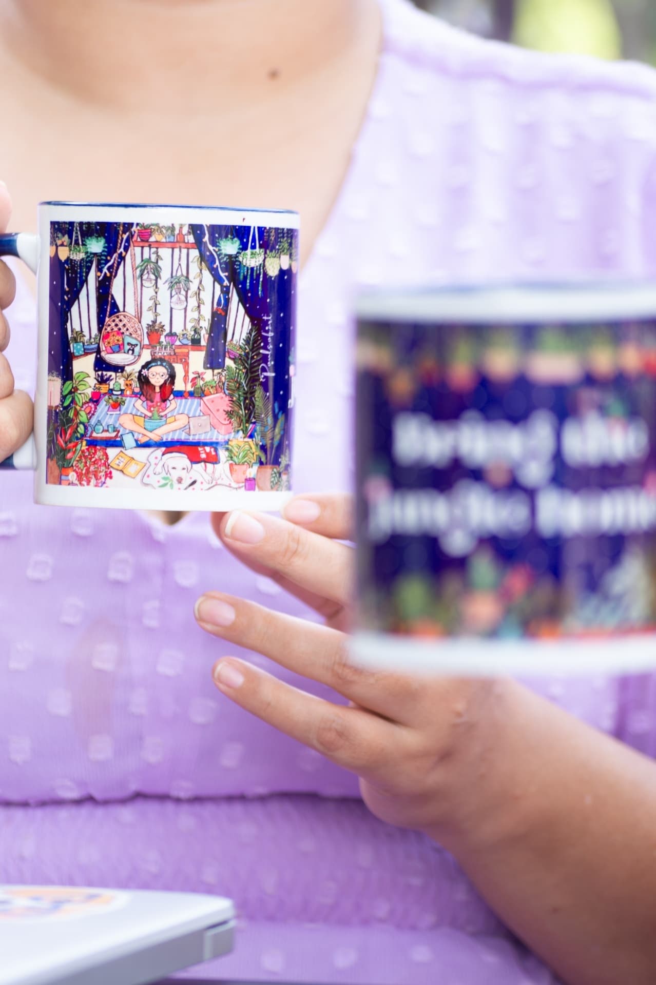 A hand holding an illustrated mug printed with a girl sitting among houseplants, with a second mug blurred in the foreground