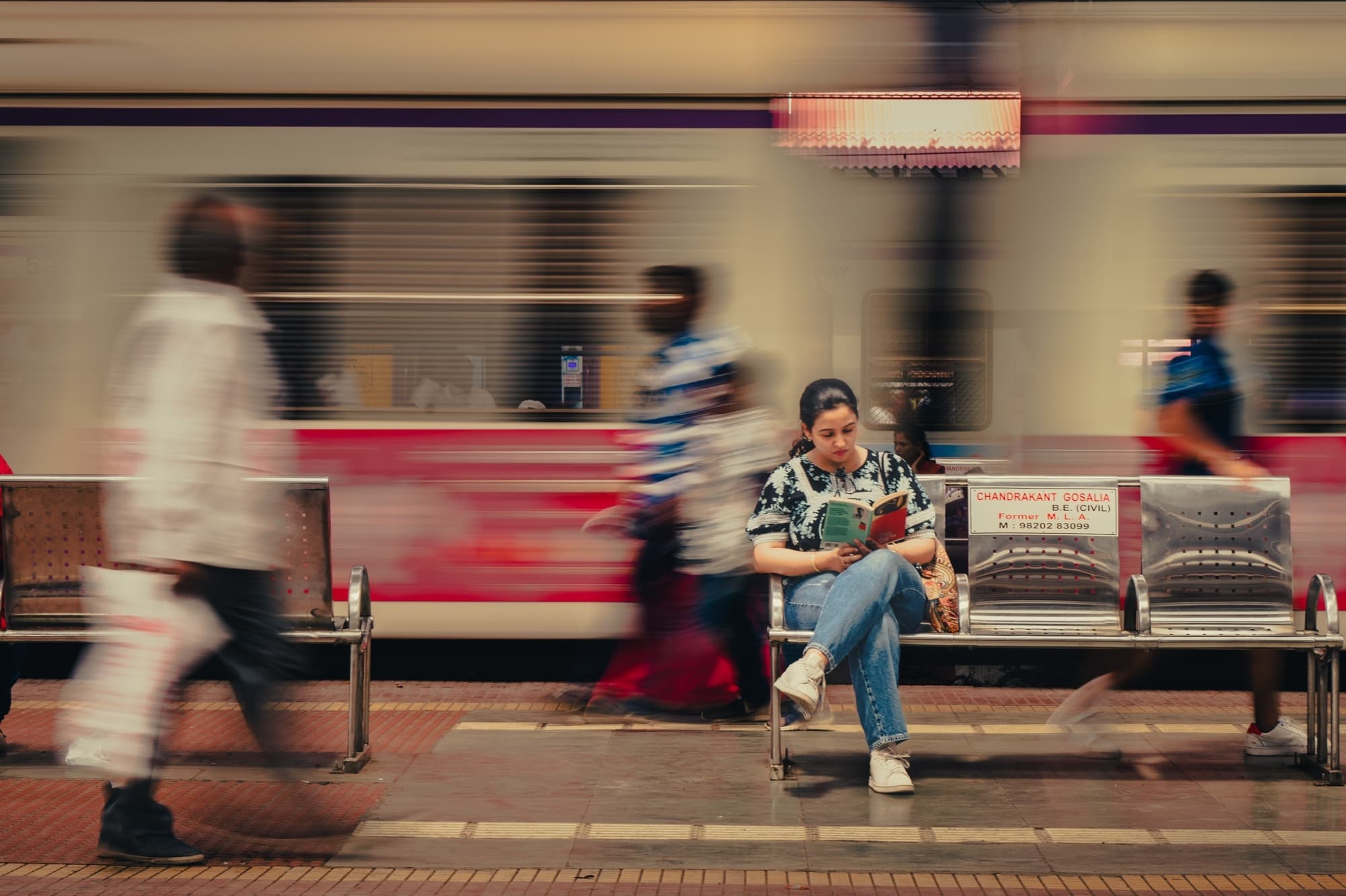 A woman reading a book on a train platform bench as a train blurs past behind her