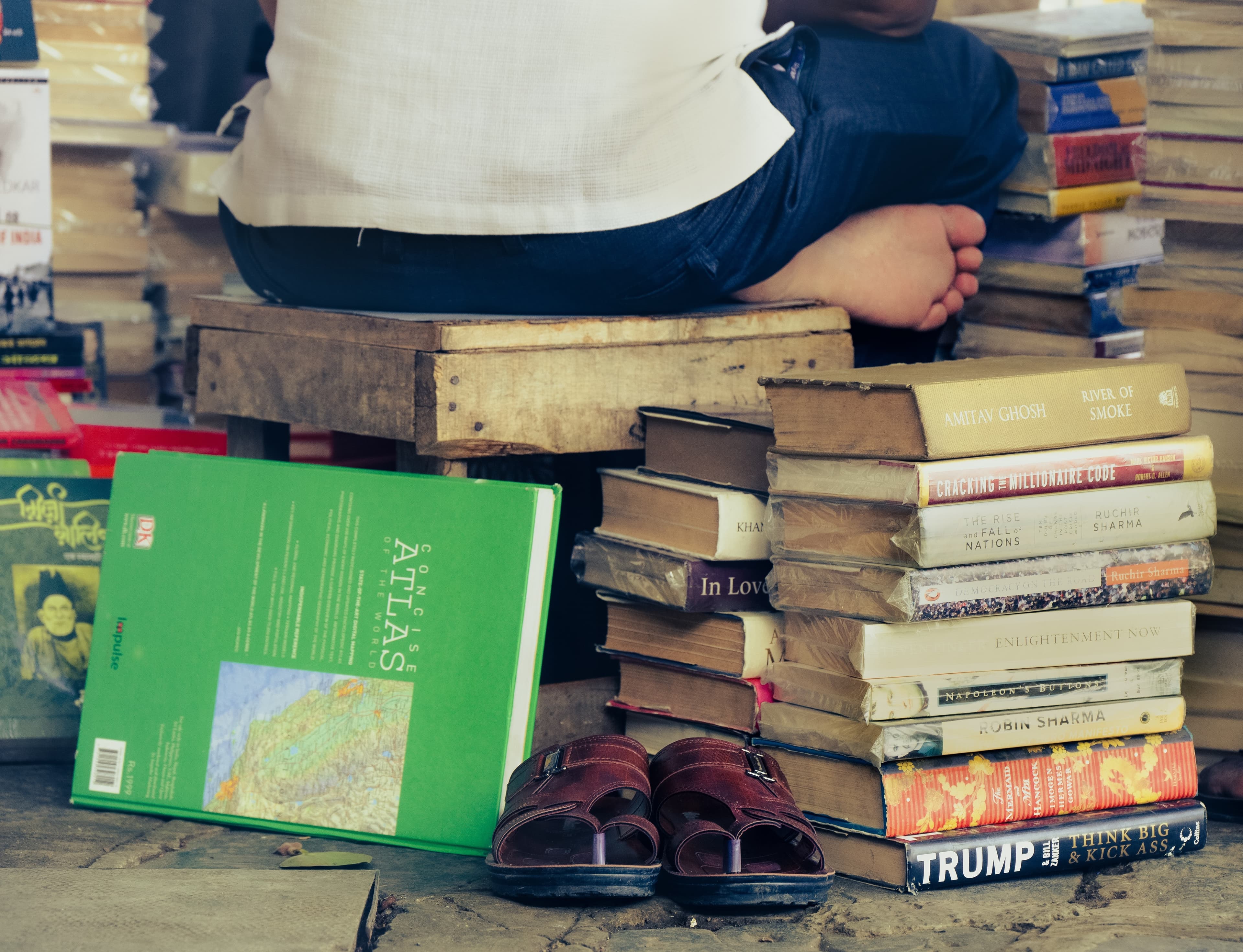 A stack of books and a pair of sandals beside a wooden stool, with someone reading behind