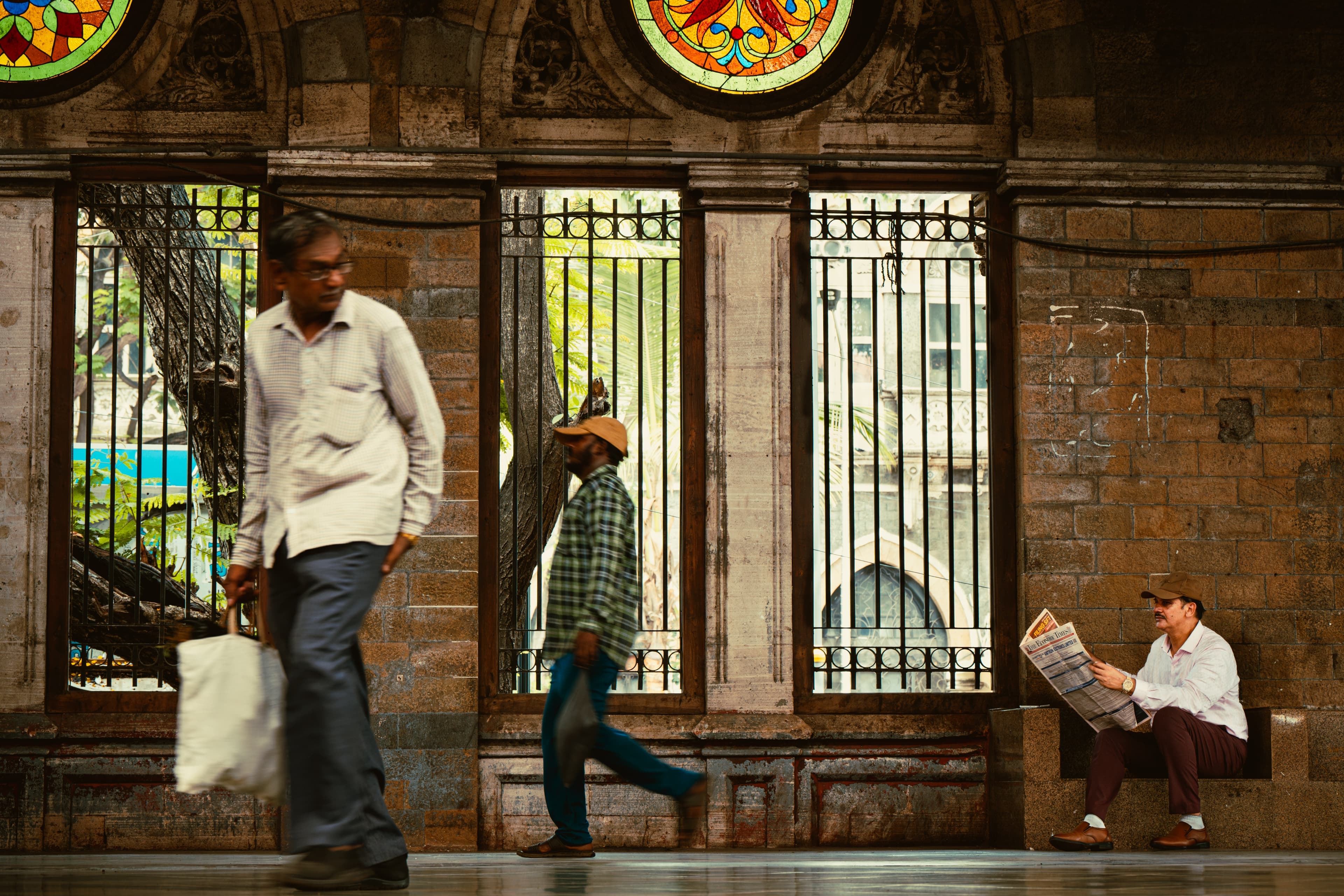 A man reading a newspaper on a bench inside a colonial-era stone gallery with stained glass windows
