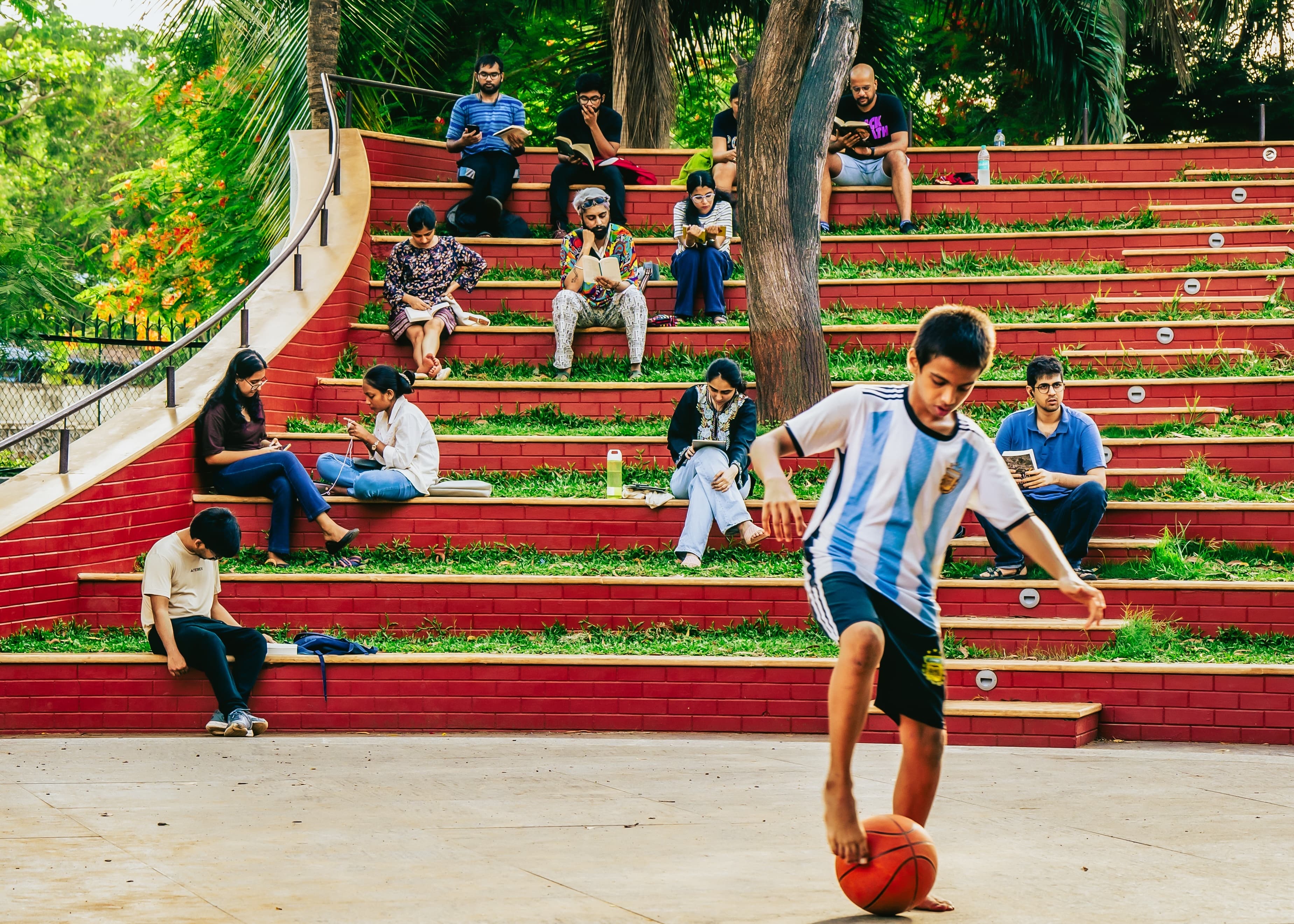 A group of people reading on red amphitheatre steps in a park, with a boy playing basketball in the foreground