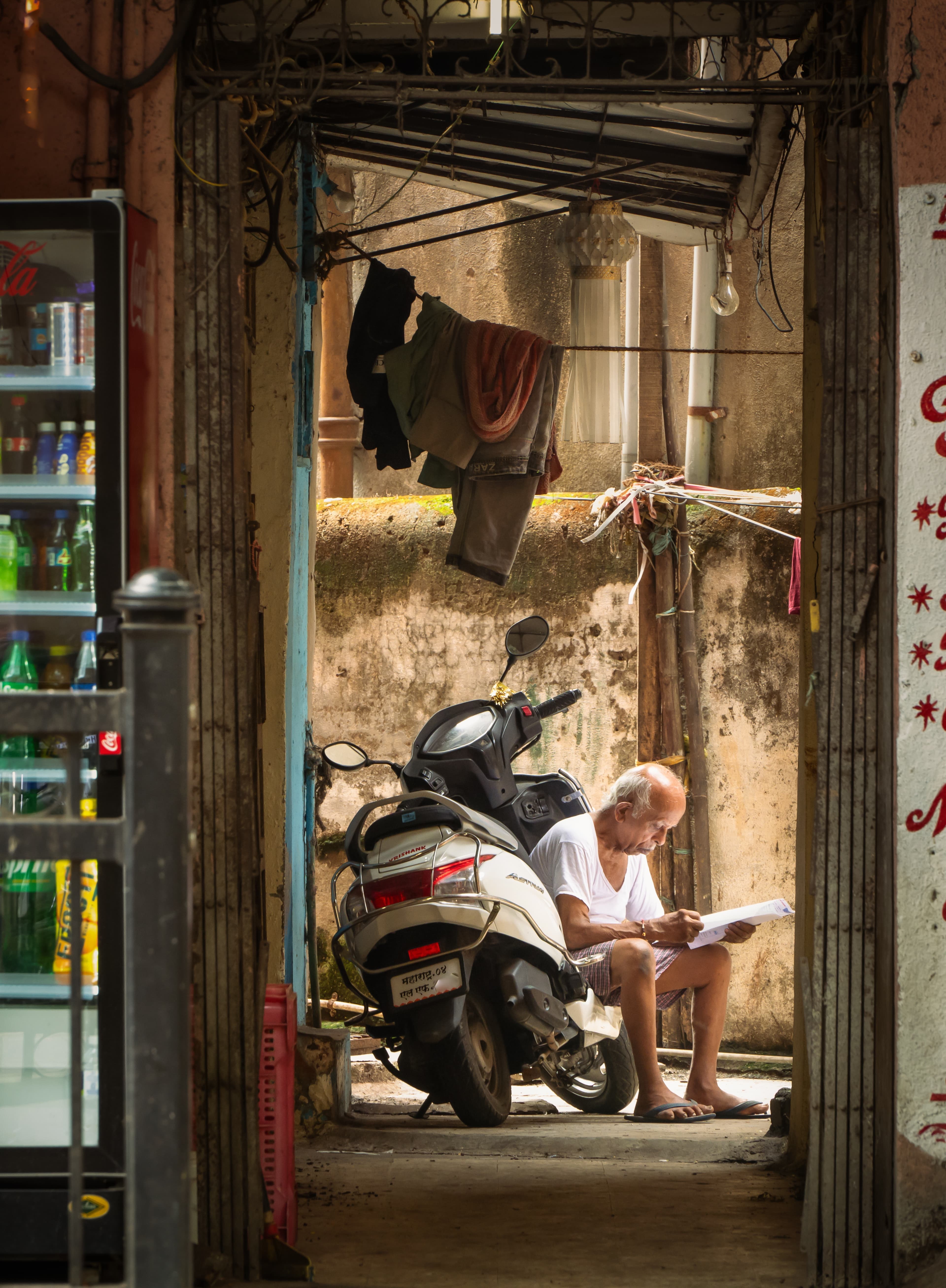 An elderly man reading papers seated beside a scooter in a shaded doorway