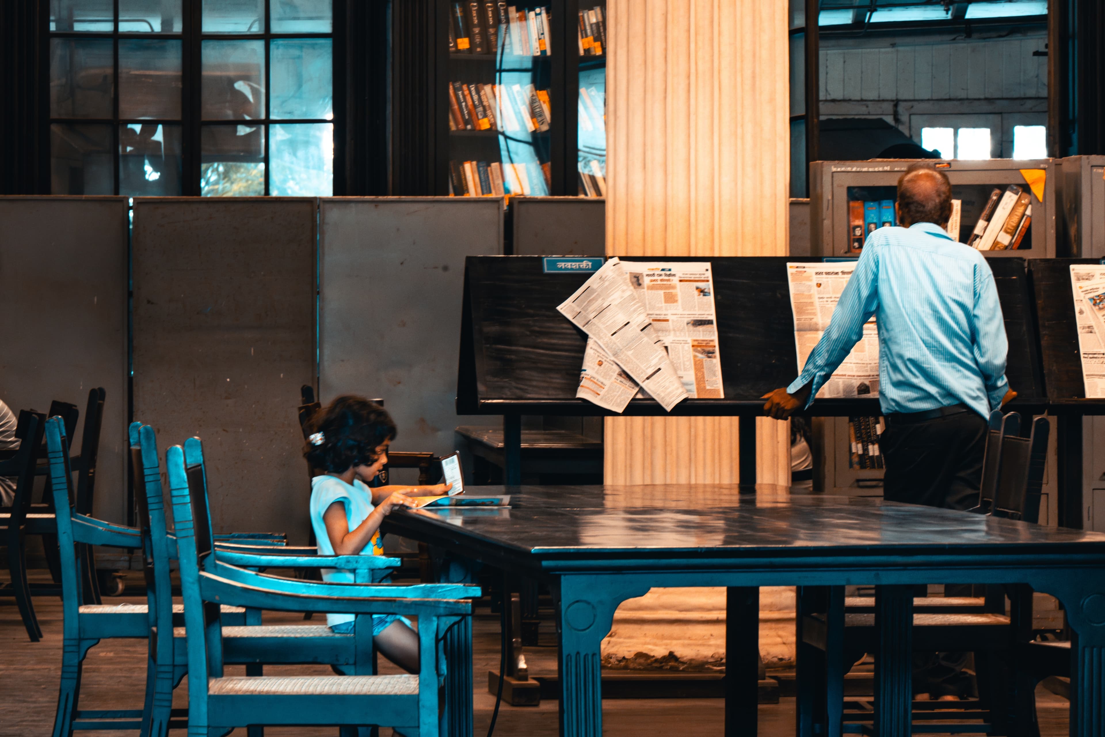 A girl reading at a table in an old library reading room, with a man browsing newspaper stands behind her