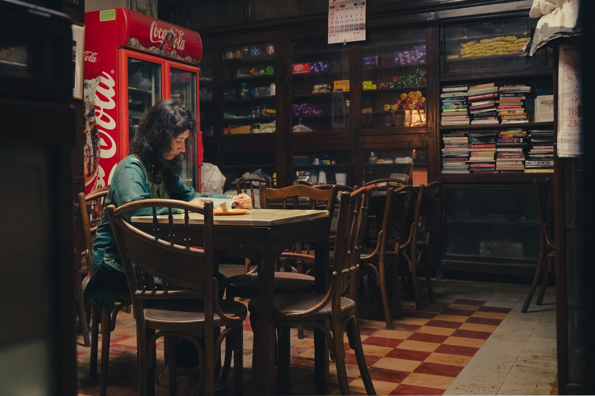 A woman reading at a table in an old-style shop with a Coca-Cola fridge and crowded bookshelves