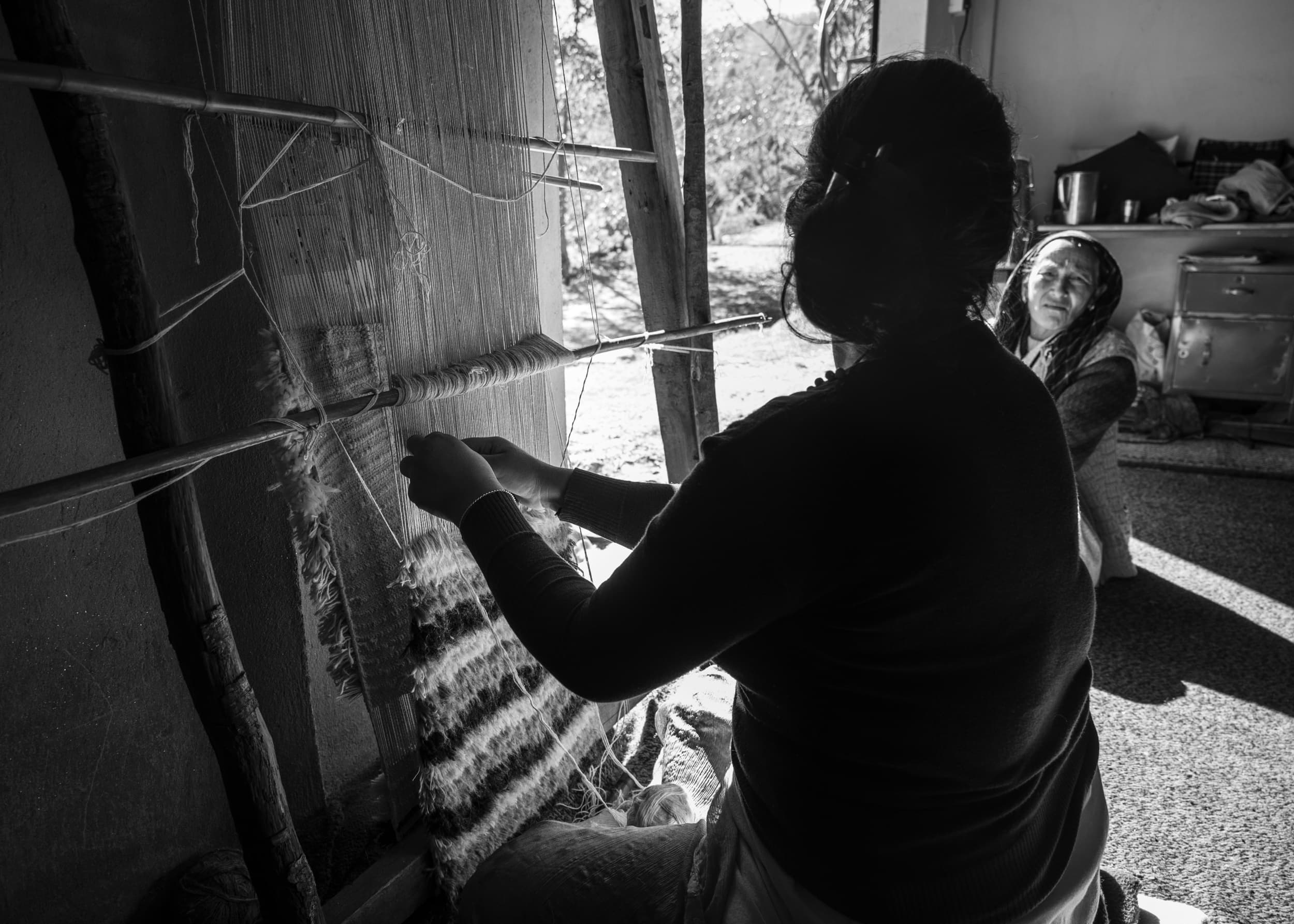 An elderly woman carding raw wool by hand indoors, with a sewing machine and framed photographs in the background, mountains visible through a window