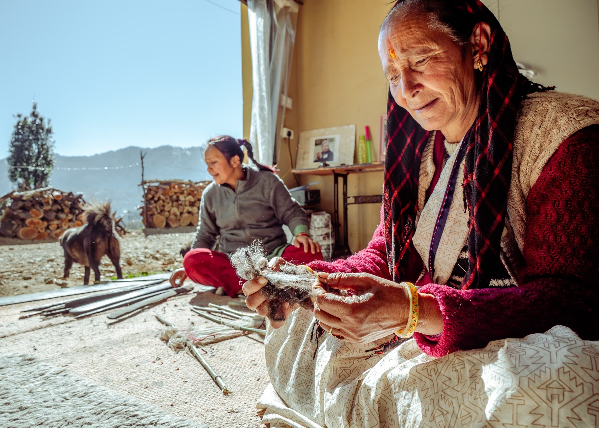 An elderly woman working with wooden loom pegs and a hammer outdoors, with stacked firewood and mountains behind her