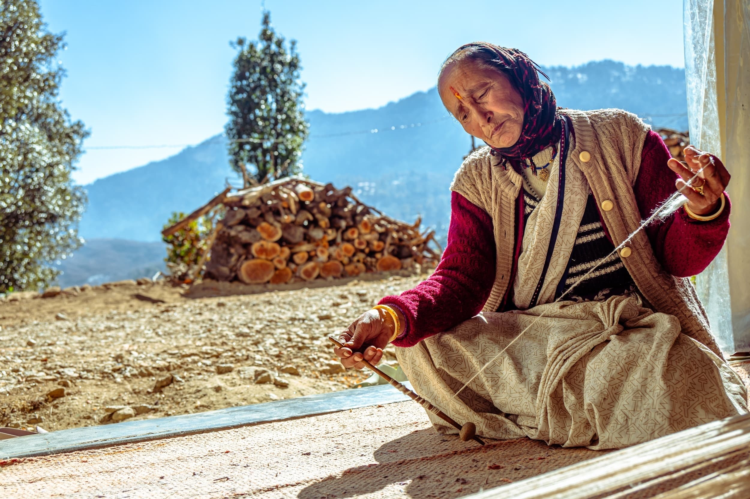 An elderly woman spinning wool with a hand-held drop spindle, seated outdoors with mountains in the distance