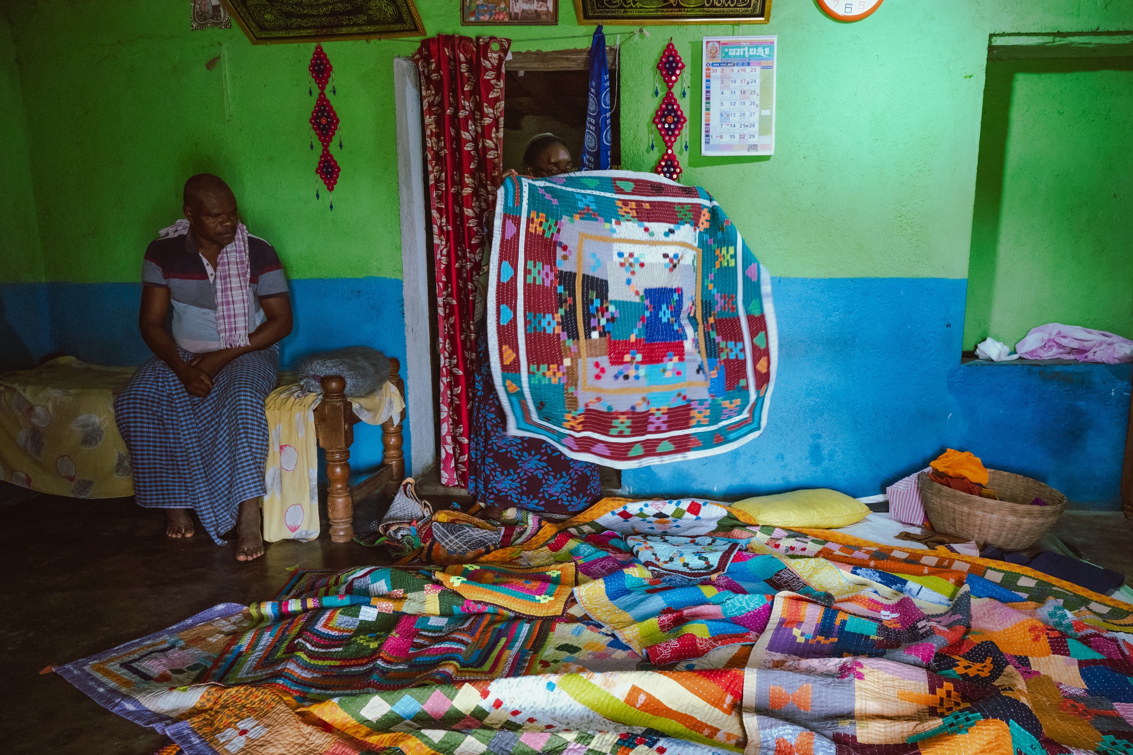 A man seated indoors as a woman peers through a curtained doorway holding up a patchwork quilt, more quilts piled on the floor
