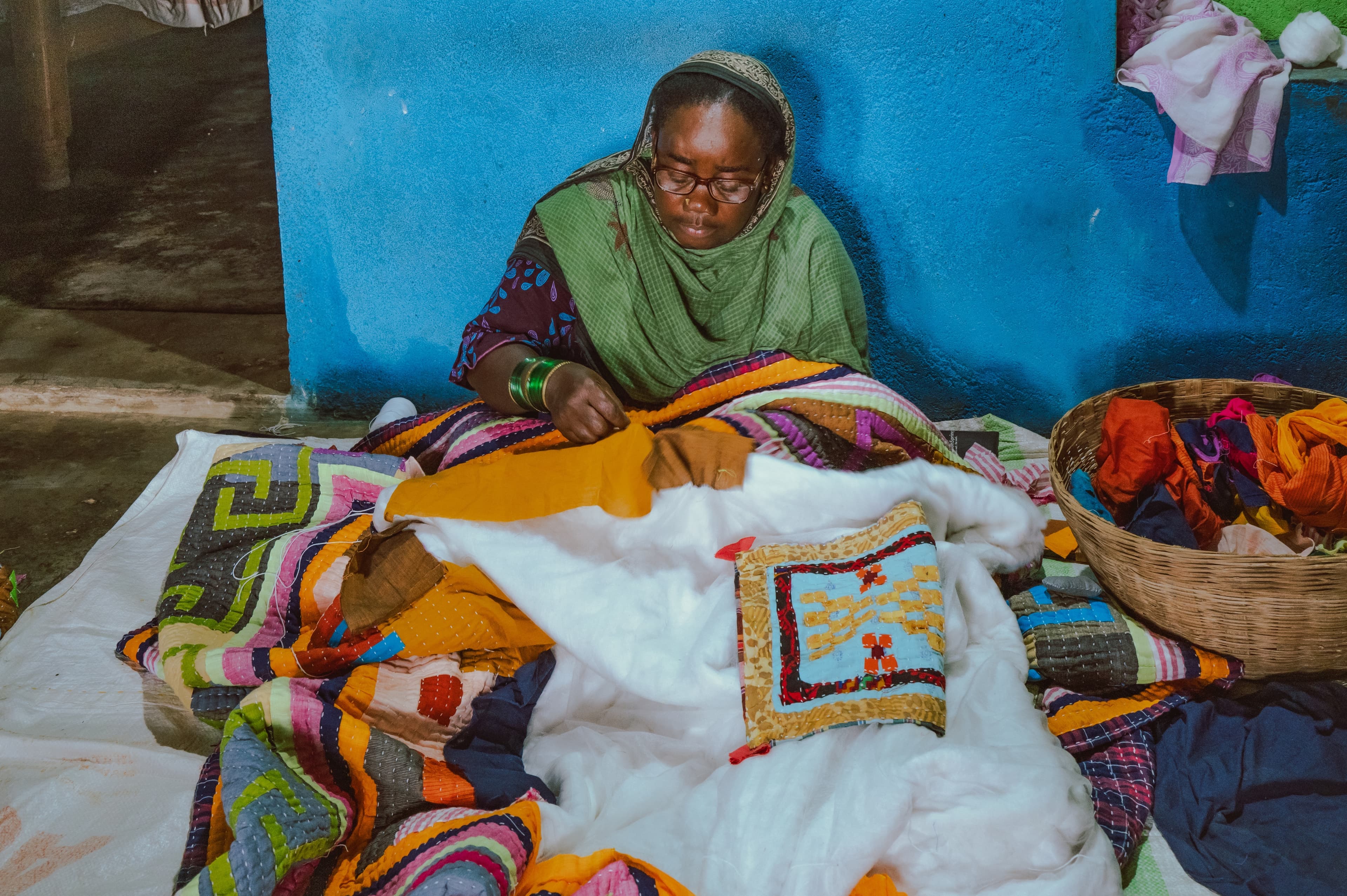 A woman in glasses hand-stitching a patchwork quilt against a blue wall, a basket of fabric scraps beside her