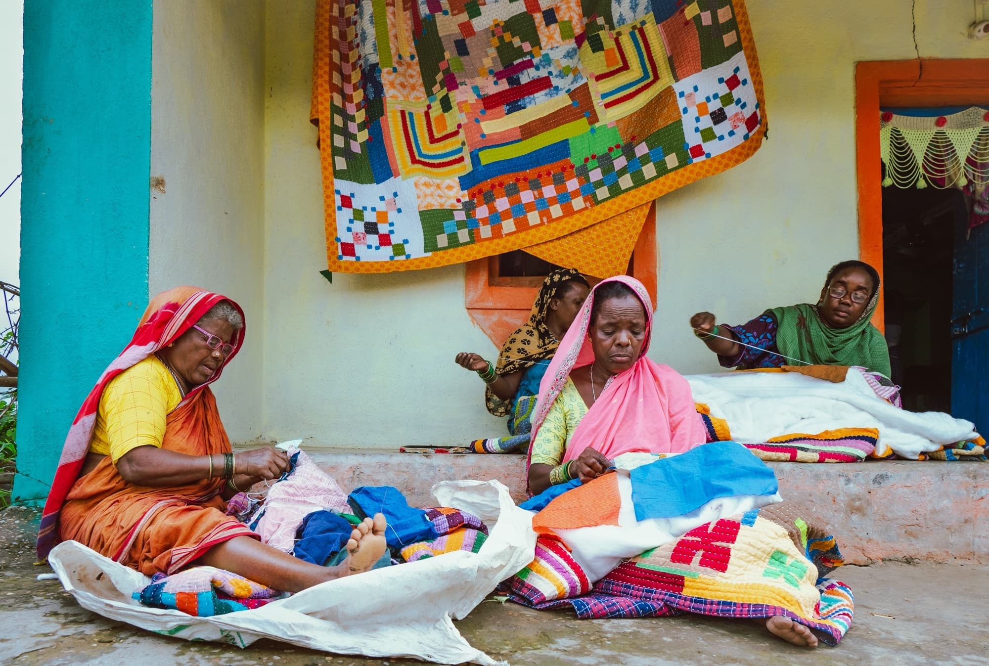 A group of women hand-stitching quilts on a porch, with a finished patchwork quilt hanging above them