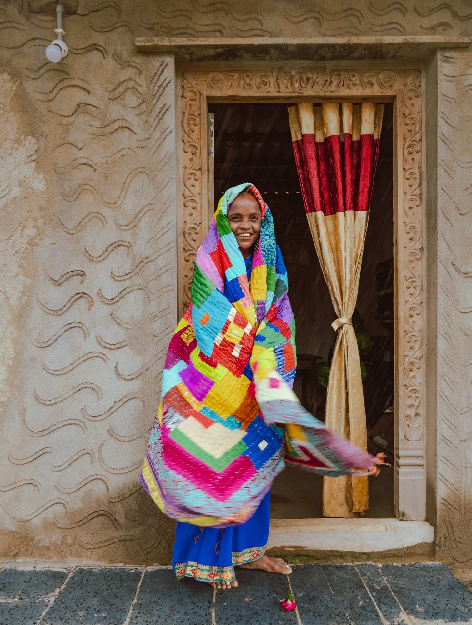 A woman twirling while wrapped in a colorful patchwork Kantha quilt in a carved stone doorway