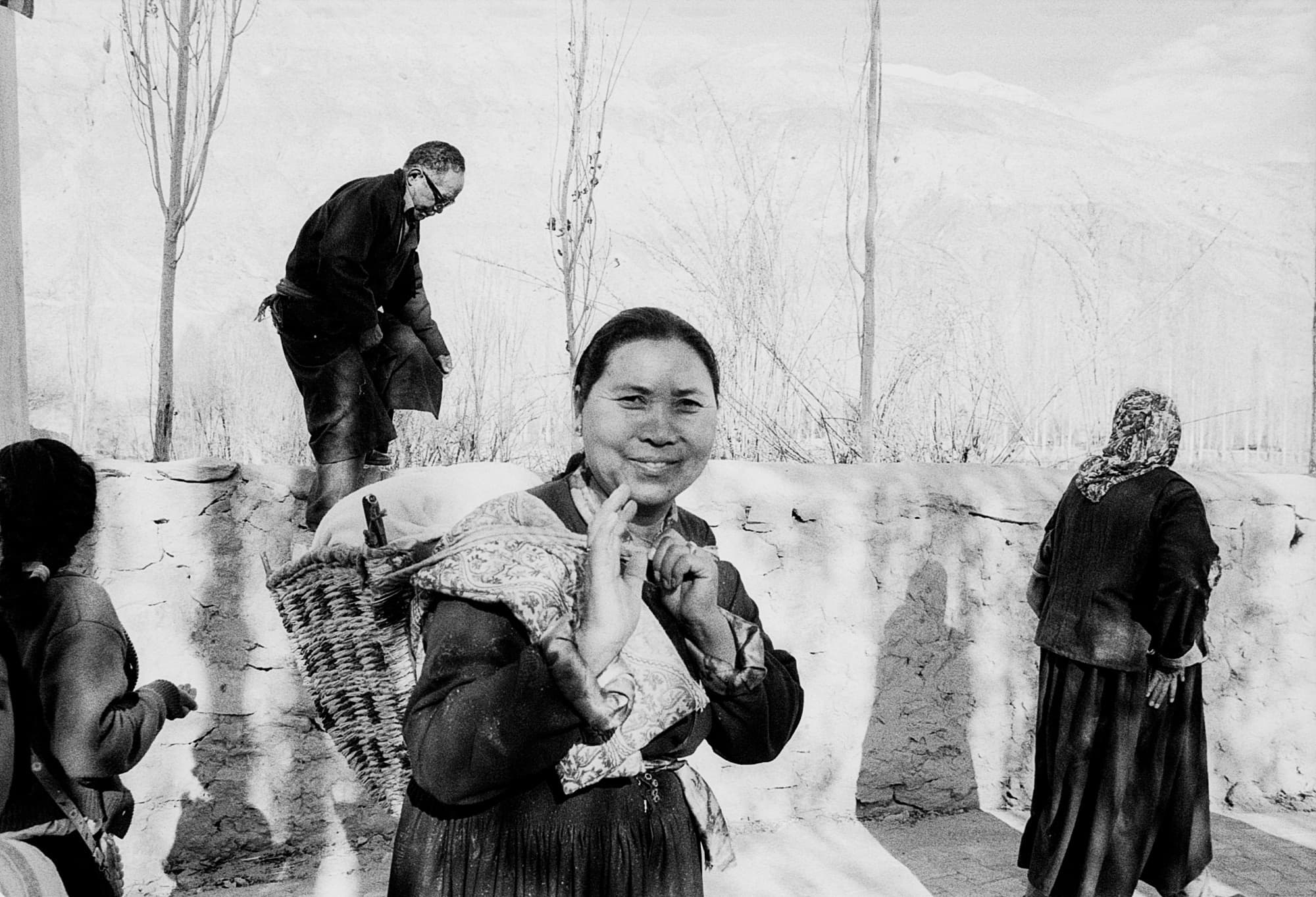 A smiling woman with a woven basket on her back, other figures and a stone wall behind her