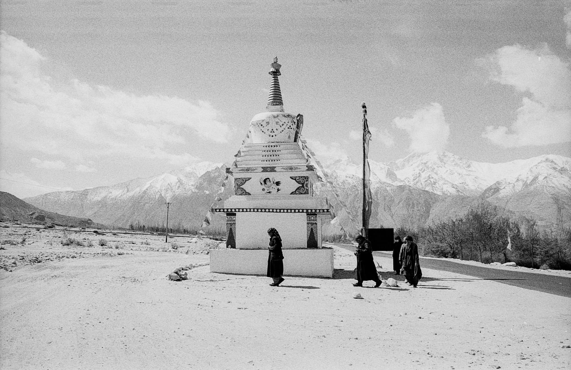 Figures walking past a whitewashed Buddhist stupa strung with prayer flags, snow-capped mountains behind