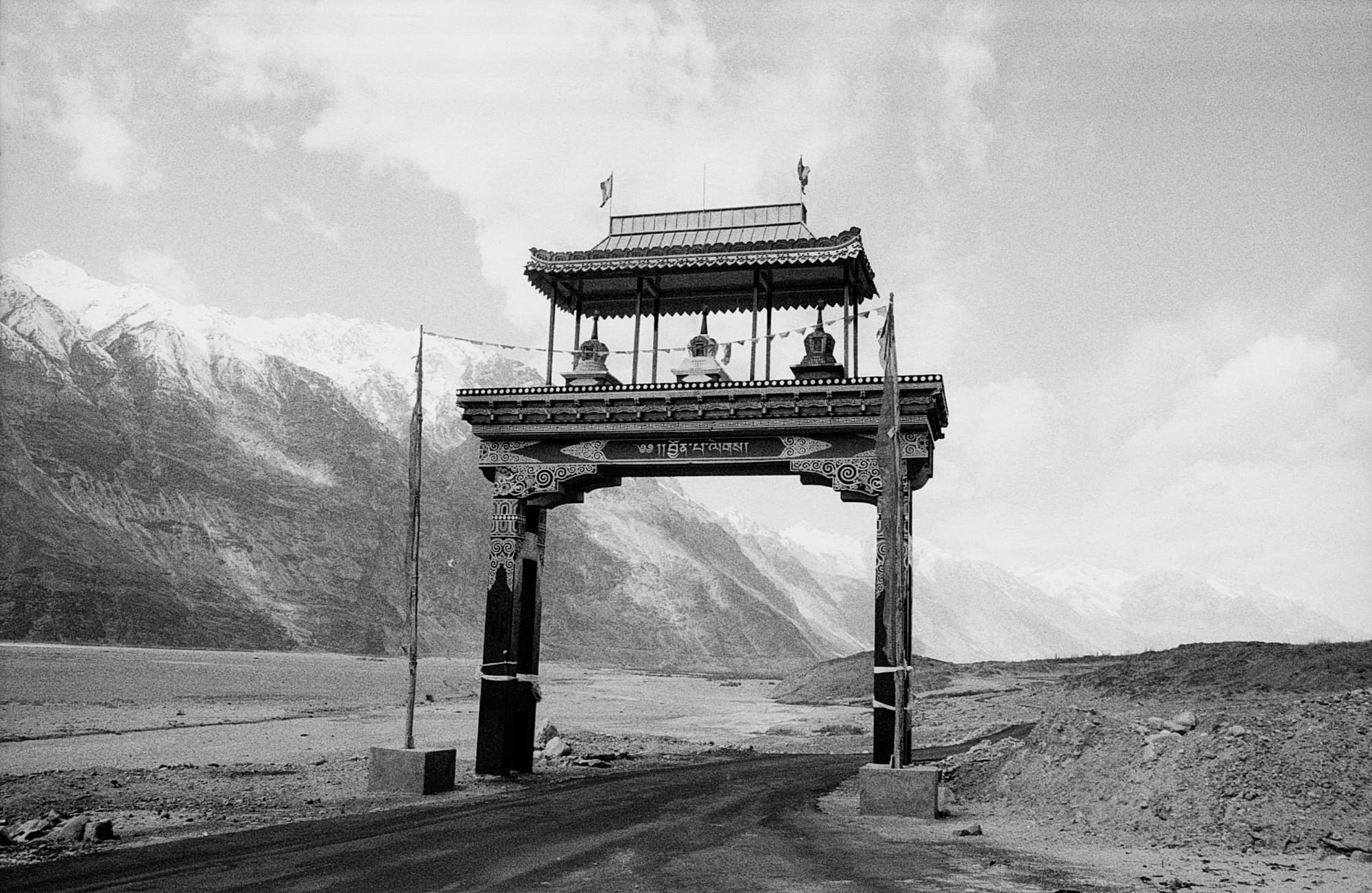An ornately decorated wooden gateway with three small stupas on top, spanning a road against a mountain valley