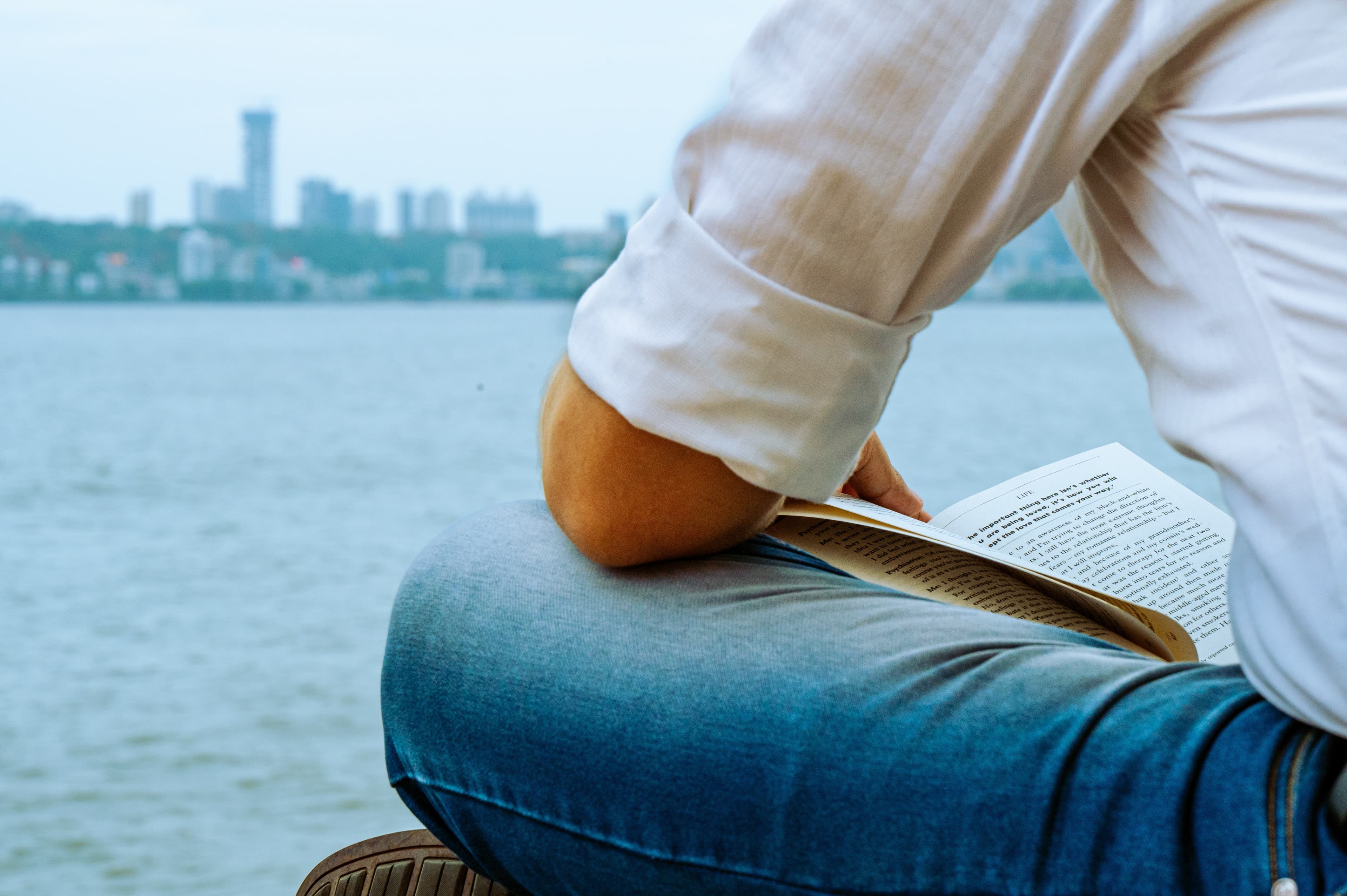 A man's arm resting on his knee while reading a paperback book, with the sea and Mumbai skyline behind him
