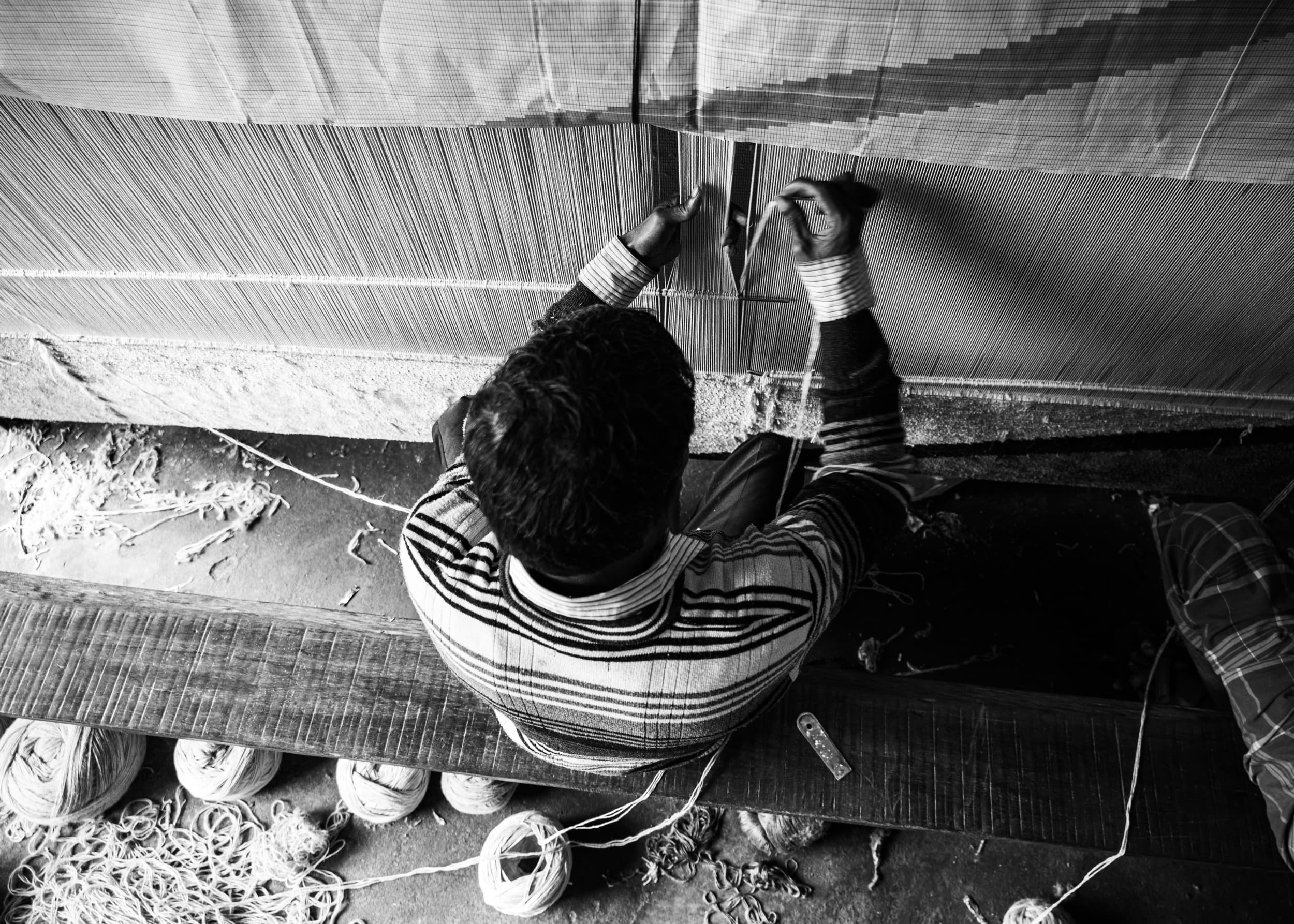An overhead view of a weaver tying knots into a carpet on a large loom, yarn balls scattered on the floor below