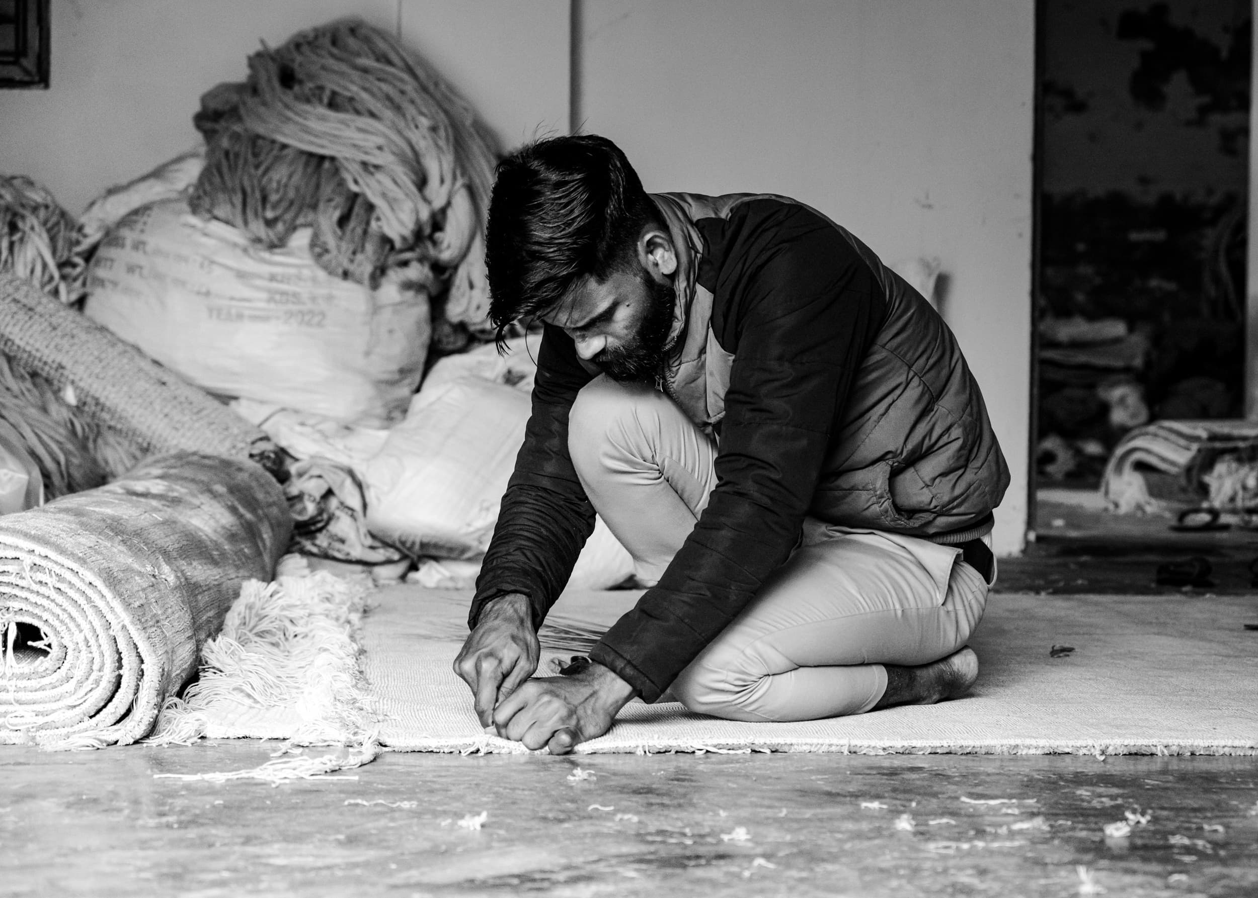 A man kneeling on the floor, trimming the edge of a rolled hand-knotted carpet
