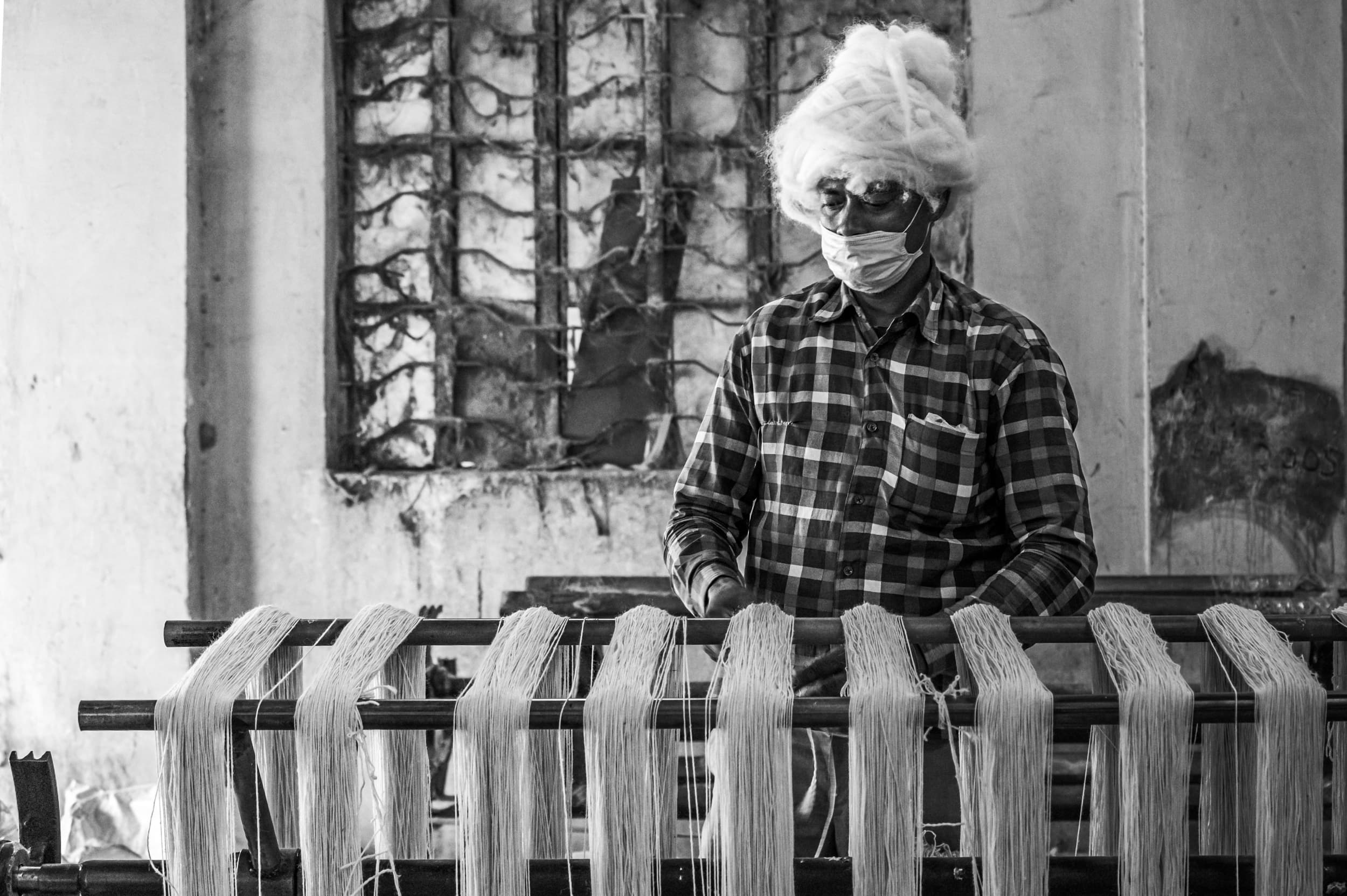 A masked worker carrying raw wool piled high on his head, standing beside a warping frame